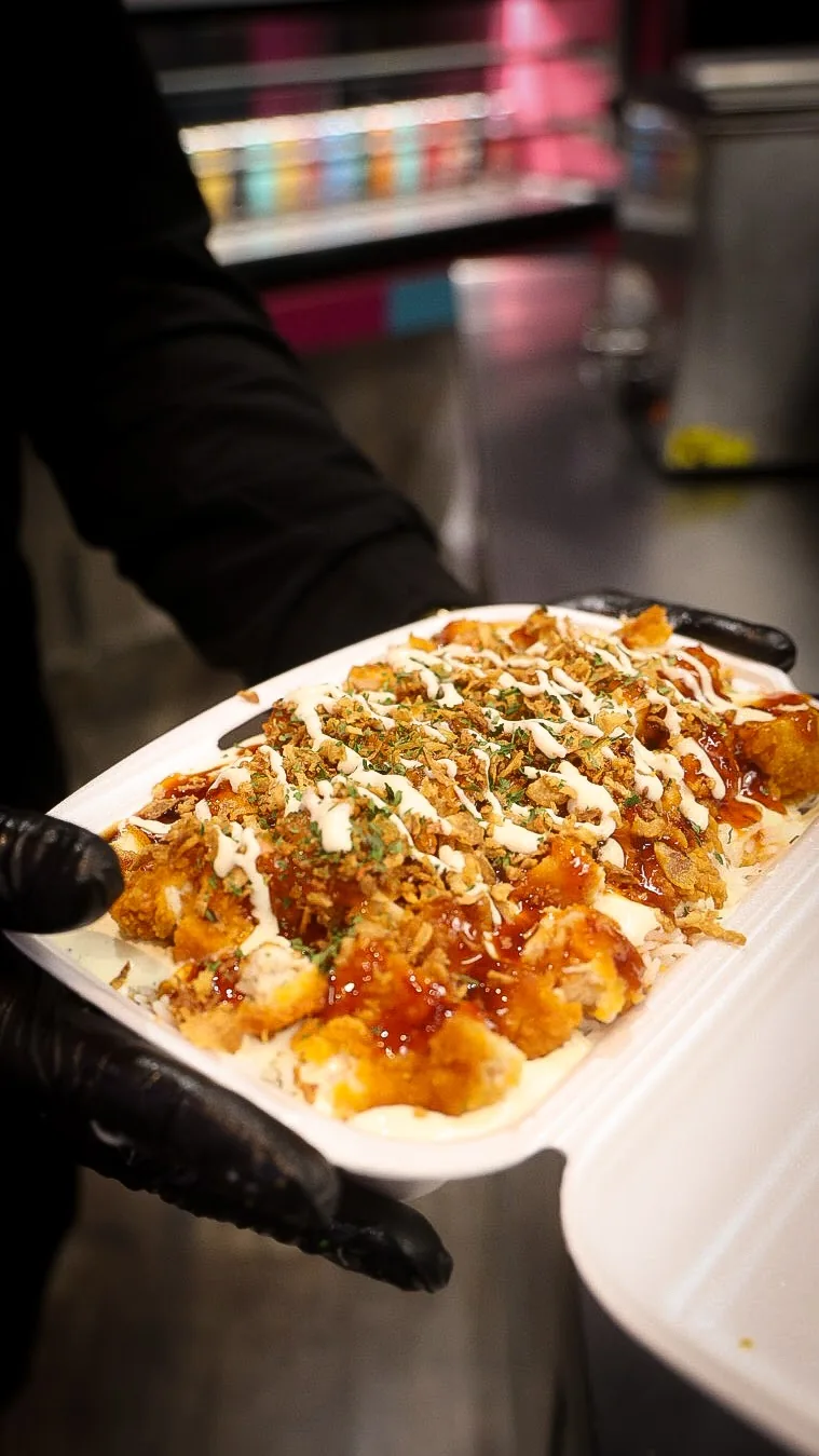 Une box Crousty Vice fraîchement préparée, tenue à la main : poulet croustillant, sauce barbecue, oignons frits et herbes sur un lit de riz